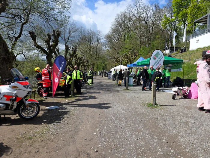 Polizeibeamte in Einsatzkleidung stehen neben Motorrädern auf einem Weg, während im Hintergrund Menschen eine Veranstaltung besuchen. Im Hintergrund sind Bäume und ein Hügel mit Zuschauertribünen zu sehen.