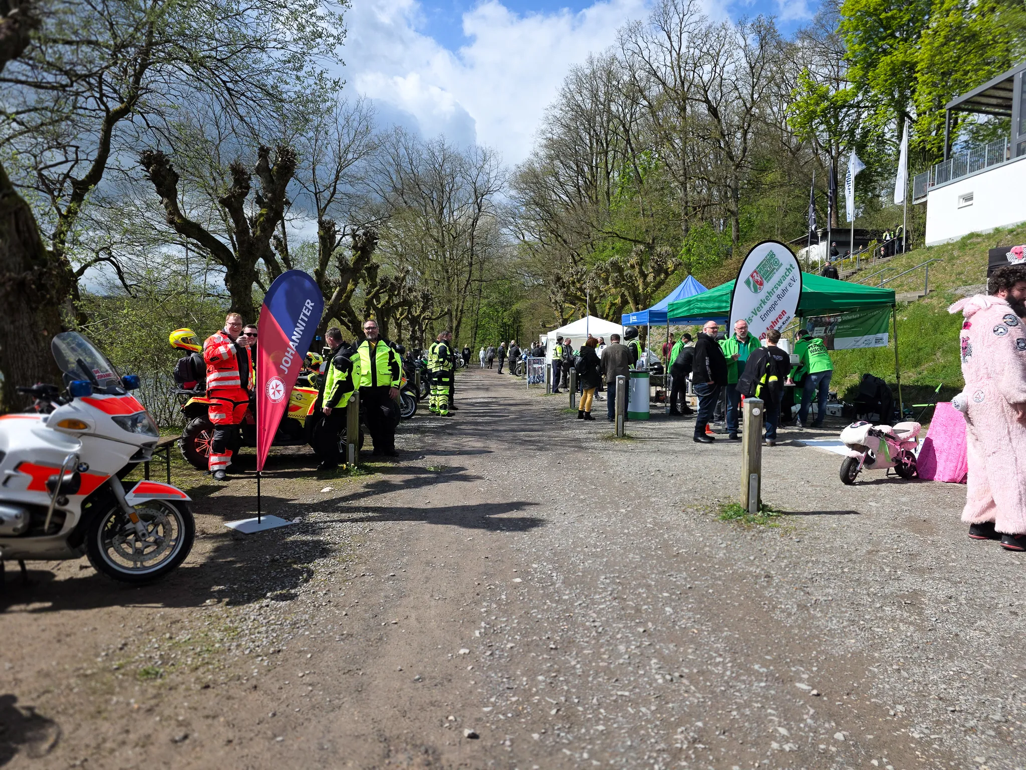 Polizeibeamte in Einsatzkleidung stehen neben Motorrädern auf einem Weg, während im Hintergrund Menschen eine Veranstaltung besuchen. Im Hintergrund sind Bäume und ein Hügel mit Zuschauertribünen zu sehen.