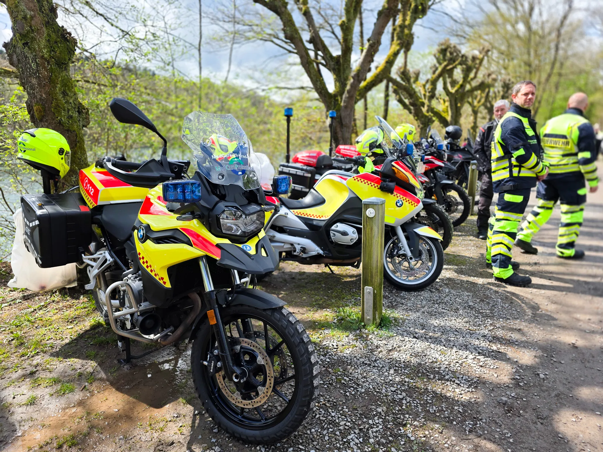 Mehrere Motorräder der Polizei mit Blaulicht und Warnstreifen stehen auf einem Parkplatz am Waldrand, während ein Polizist im Gespräch ist.