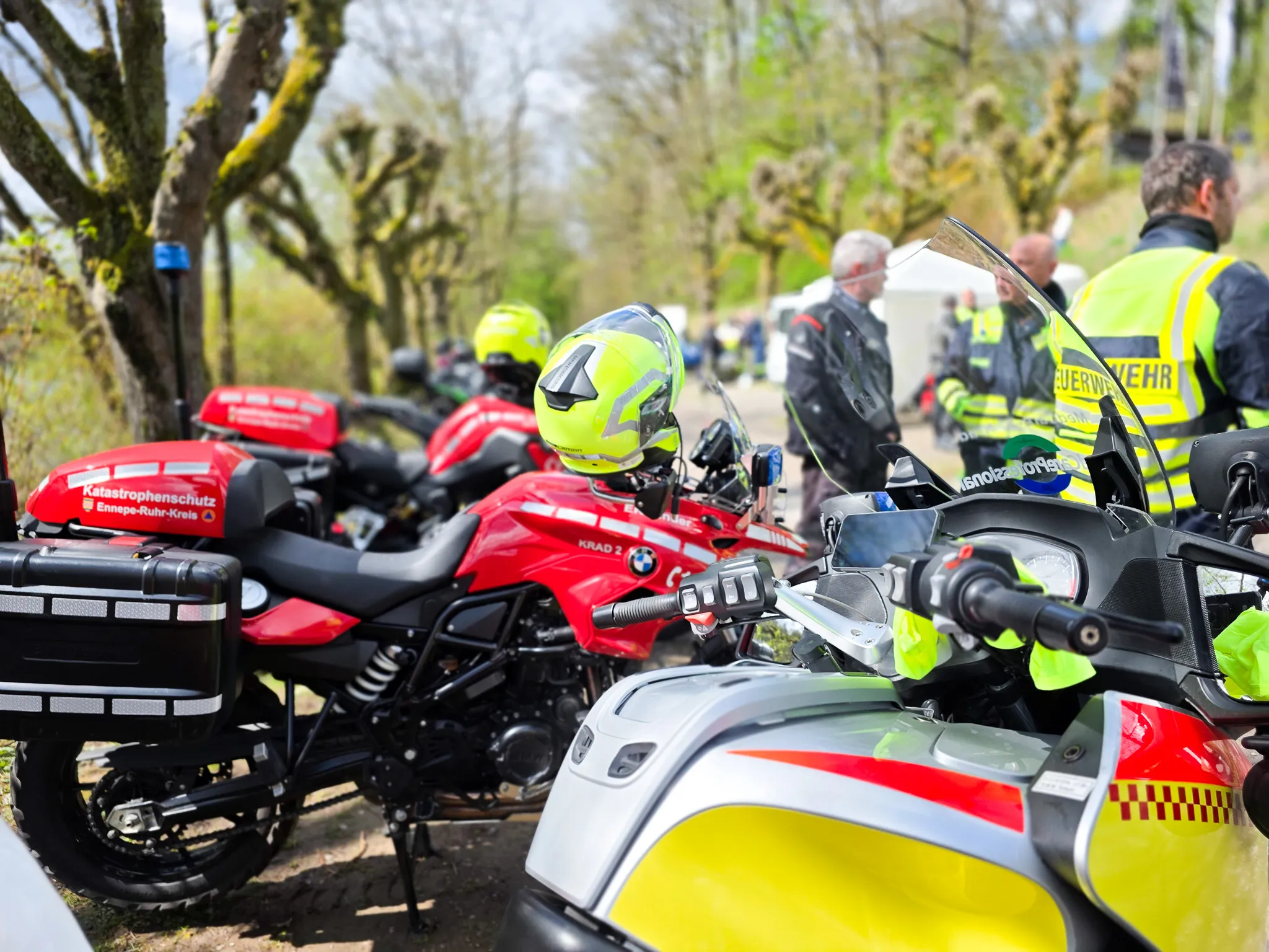 Mehrere Motorräder der Polizei und des Katastrophenschutzes stehen auf einem Parkplatz, im Hintergrund sind Einsatzkräfte zu sehen. Die Motorräder sind rot und weiß und mit Warnstreifen versehen.