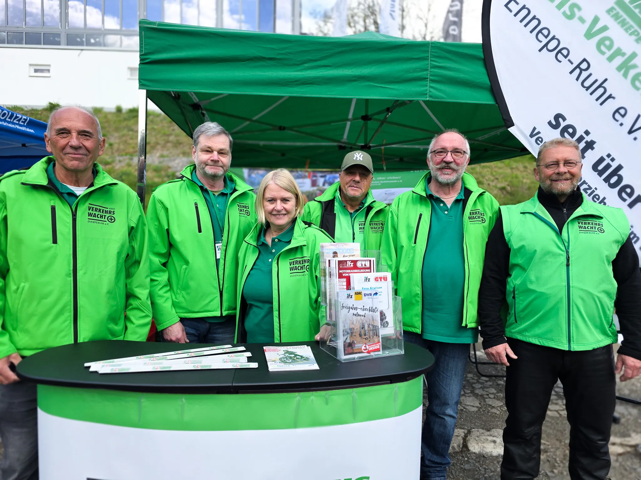 Mehrere Personen in grünen Jacken mit dem Aufdruck „Verkehrswacht“ stehen an einem Infostand unter einem Zelt. Im Hintergrund ein Banner der Polizei und Verkehrssicherheitsempfehlungen für Motorradfahrer.