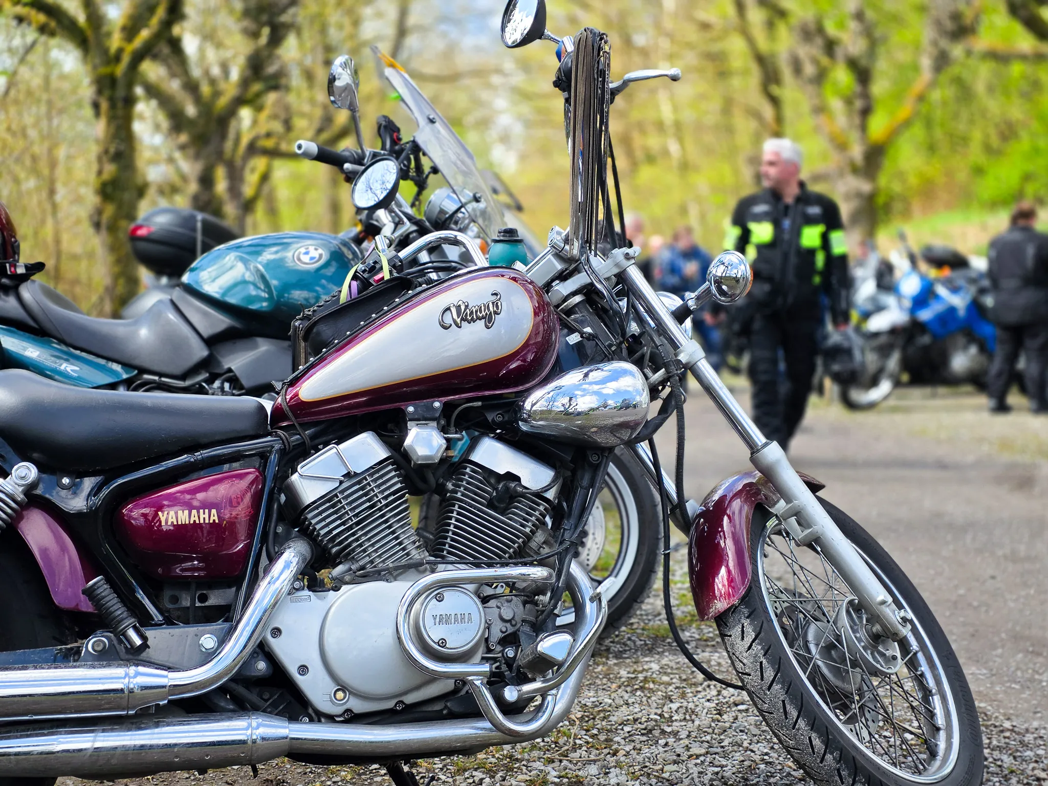 Mehrere geparkte Motorräder, darunter ein dunkelrotes Yamaha-Modell im Vordergrund, stehen auf einem Kiesplatz. Im Hintergrund sind Polizisten und weitere Motorradfahrer zu sehen.