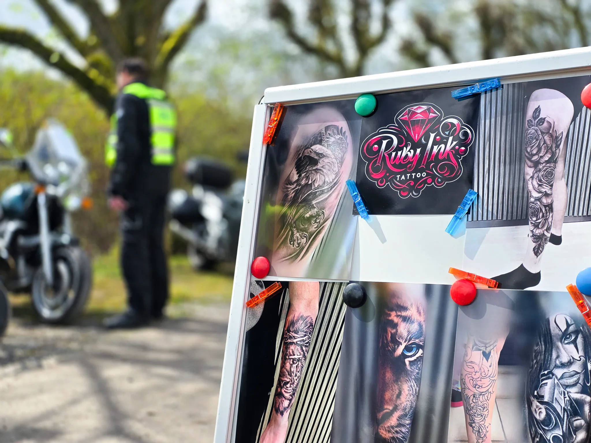 Werbetafel für „Ruby Ink Tattoo“ mit verschiedenen Tattoo-Designs im Hintergrund einer Präventionsveranstaltung der Polizei für Biker.