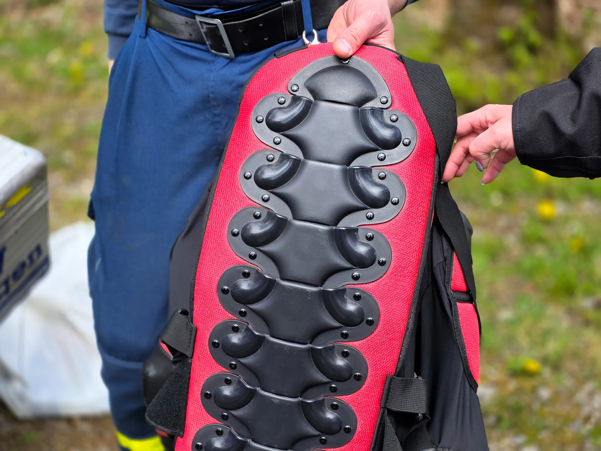 Roter Motorrad-Rückenprotektor mit schwarzen Polstern und Schnallen, der von einer Person gehalten wird. Im Hintergrund ist ein Teil einer Polizeiuniform zu sehen.