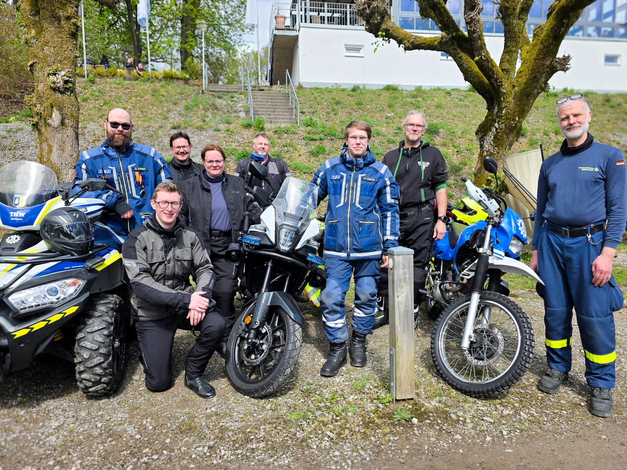 Eine Gruppe von Polizisten in Motorradmontur steht mit zwei Motorrädern vor einer Grünfläche. Im Hintergrund ein Gebäude mit einer Treppe und Bäume.