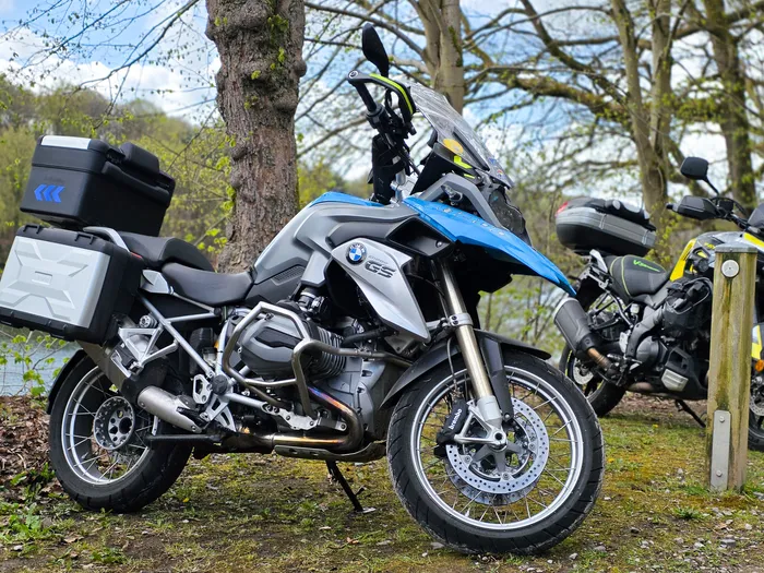 Blaue BMW-Motorrad mit Gepäckträgern steht am Ufer eines Sees neben einem weiteren Motorrad, im Hintergrund Bäume und ein bewölkter Himmel.