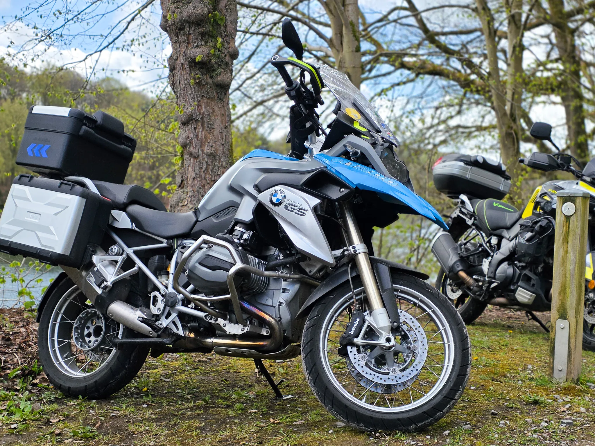 Blaue BMW-Motorrad mit Gepäckträgern steht am Ufer eines Sees neben einem weiteren Motorrad, im Hintergrund Bäume und ein bewölkter Himmel.