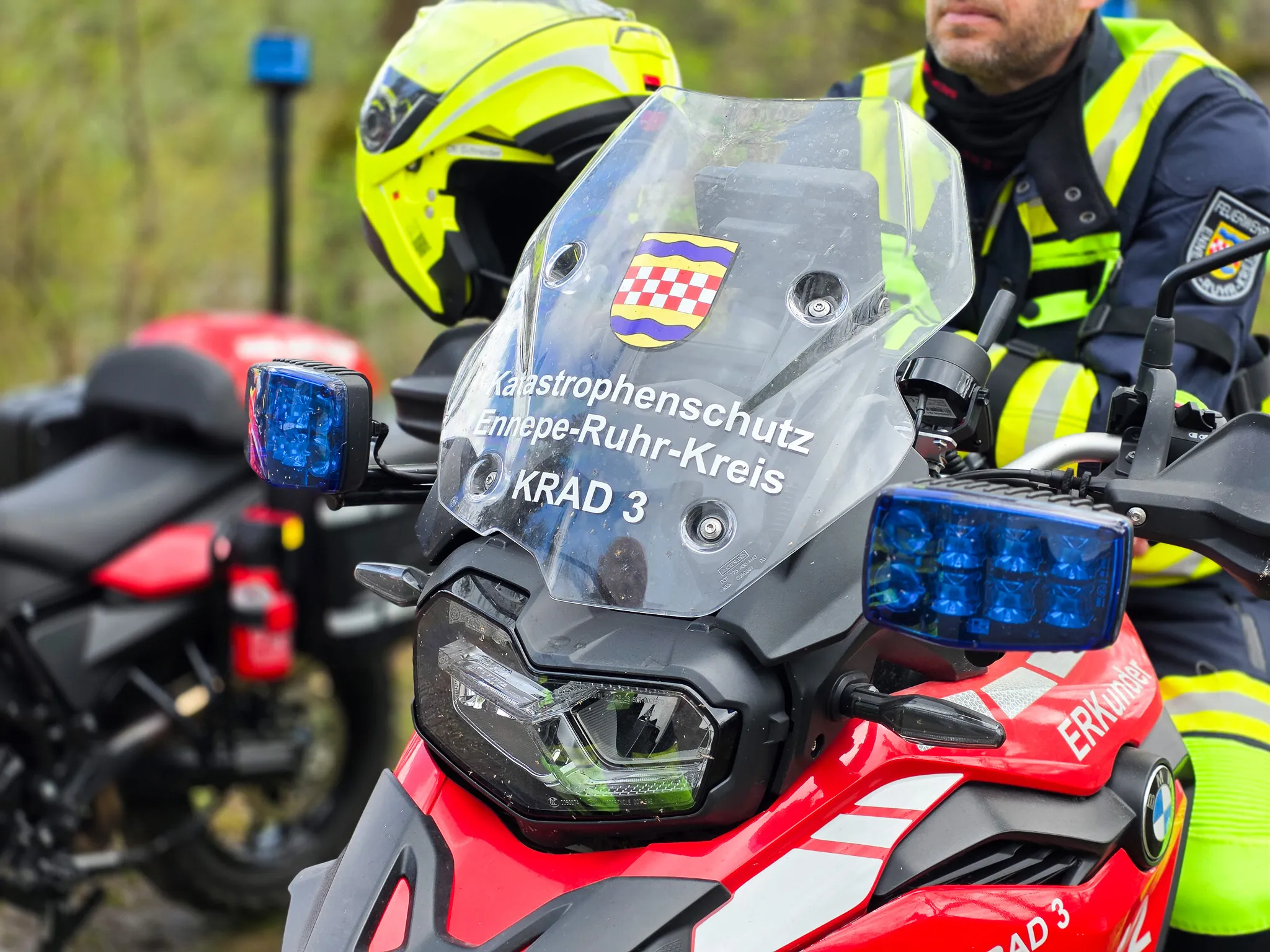 Motorrad des Katastrophenschutzes Ennepe-Ruhr-Kreis mit Blaulicht und Aufschrift „KRAD 3“ steht im Fokus, im Hintergrund ein Polizist mit Helm.