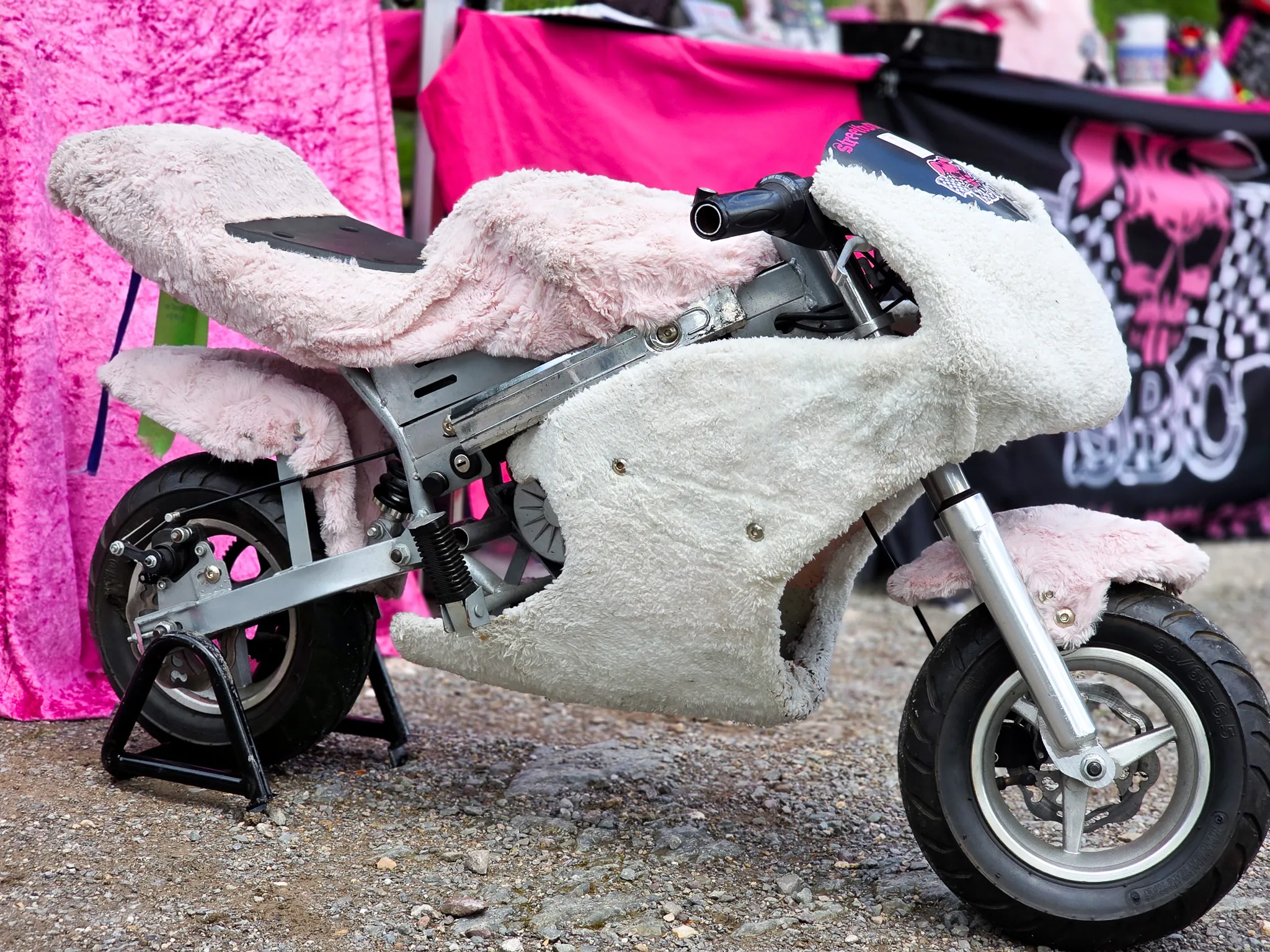 Kleines Motorrad mit flauschigem, weißem Überzug steht auf einem Kiesweg vor einem Stand mit Rennflaggen.