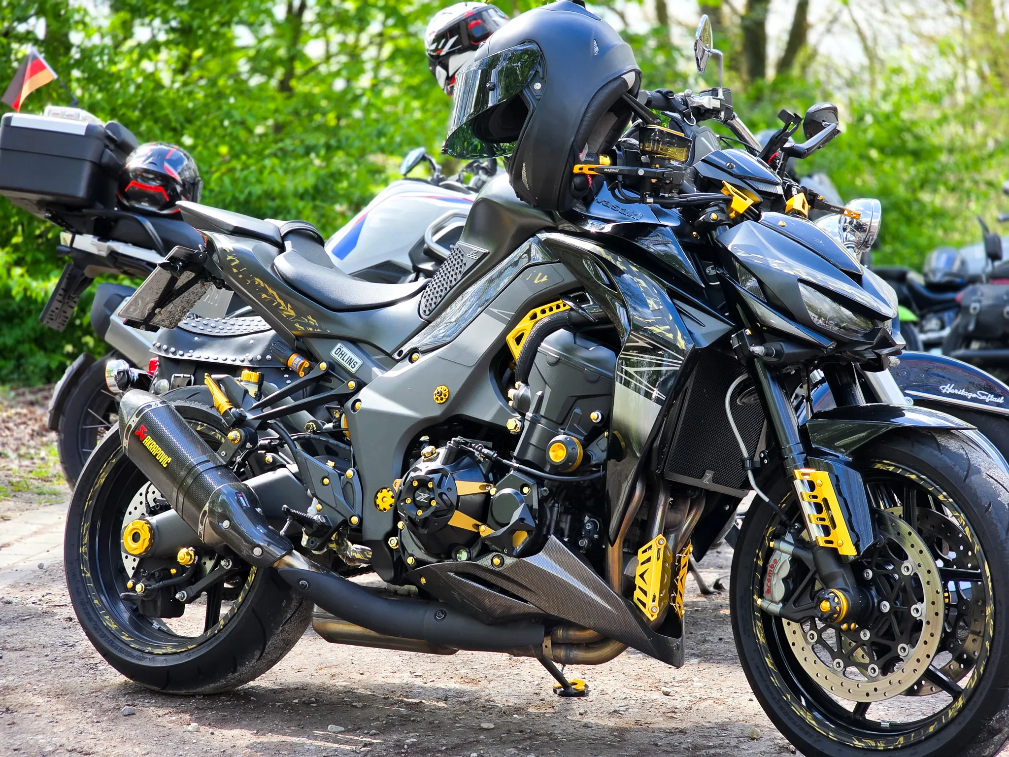 Dunkelgraues Motorrad mit auffälligen goldenen Akzenten und einem schwarzen Helm auf dem Sitz steht auf einem Kiesweg. Im Hintergrund sind weitere Motorräder und Bäume zu sehen.