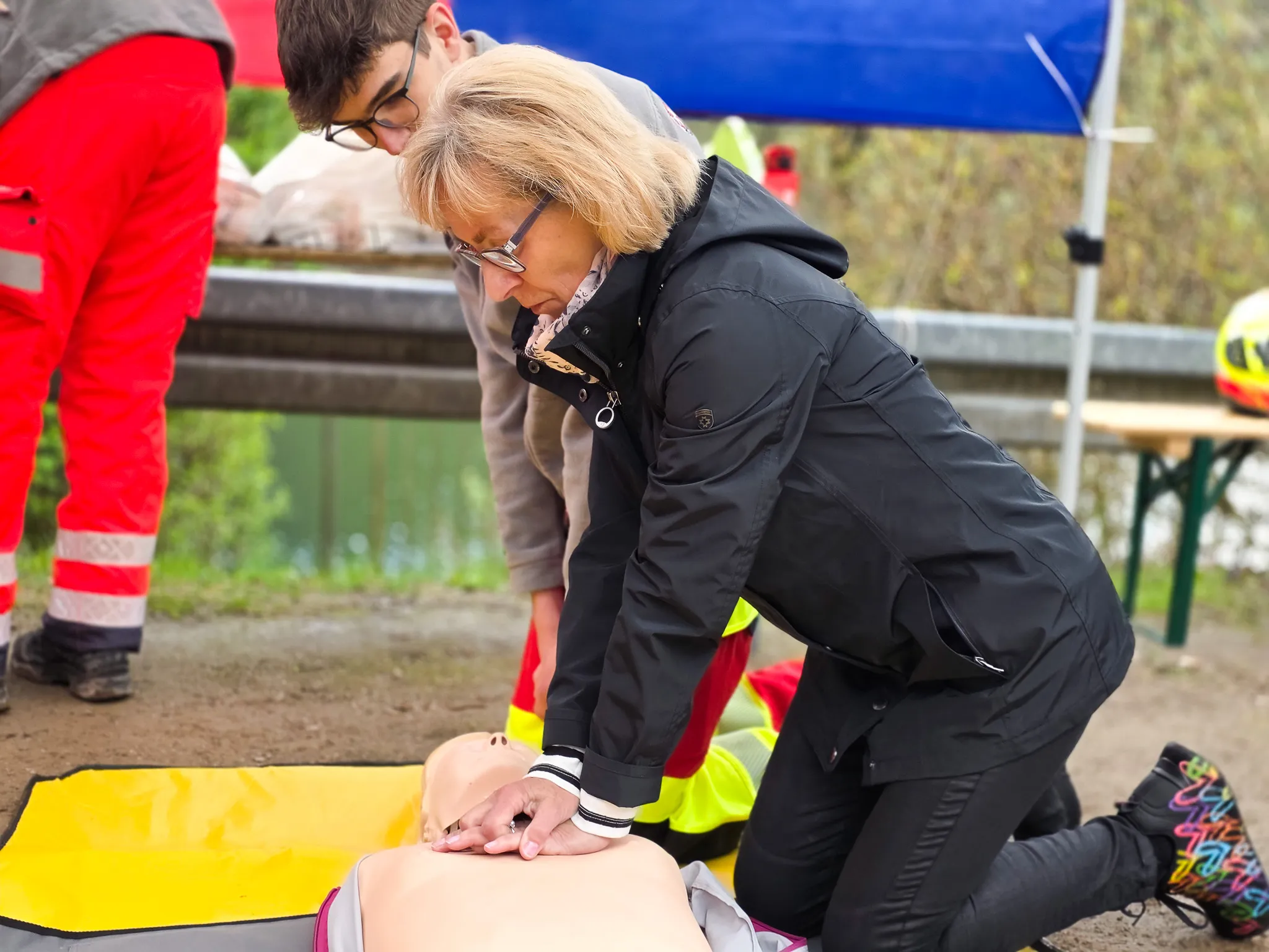Eine Frau in einem schwarzen Jackett führt an einer Person auf einer gelben Matte Herzdruckmassage durch, während ein Mann im Hintergrund zusieht.