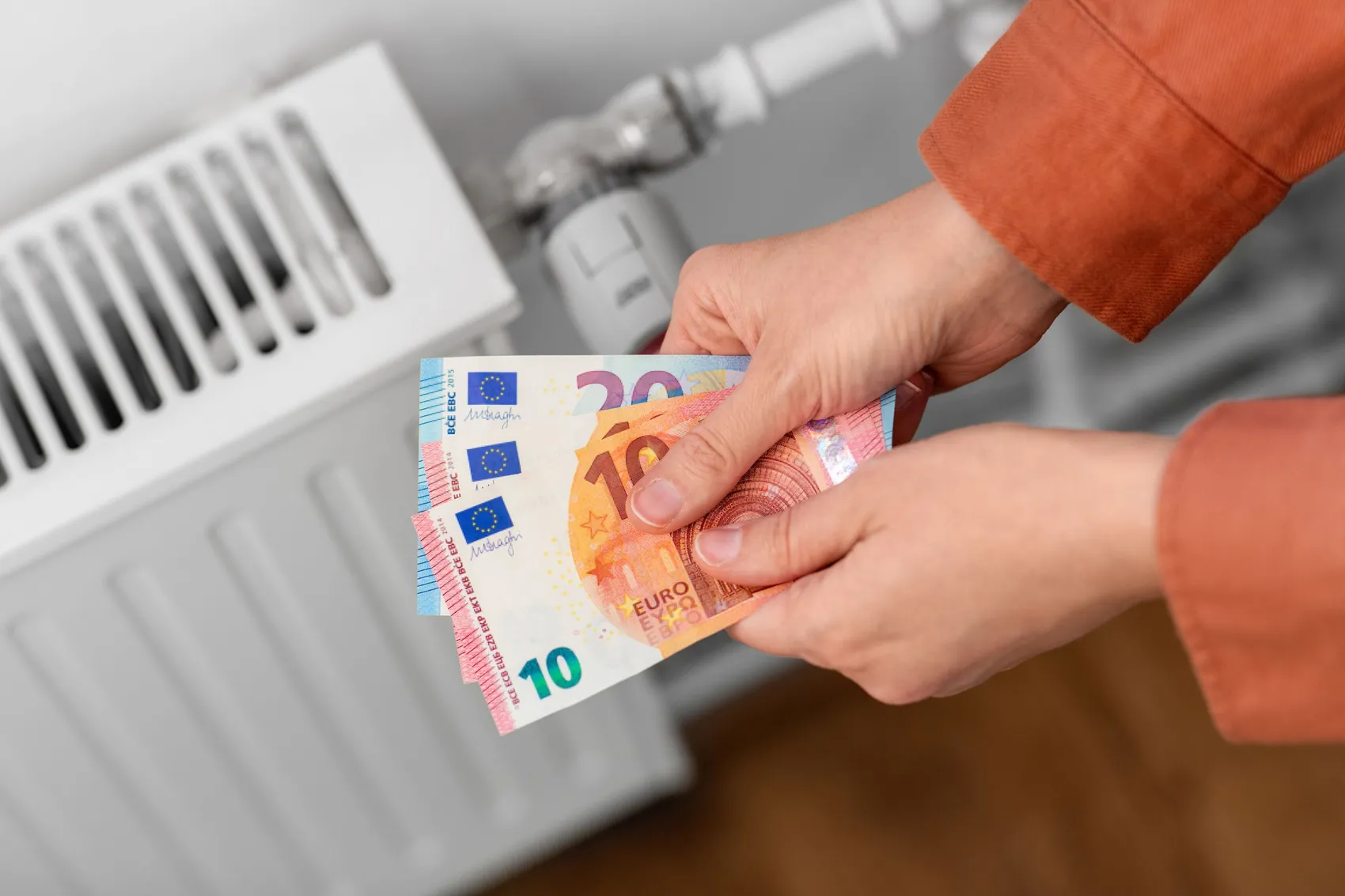 Eine Hand hält Euro-Banknoten vor einem Heizkörper, was steigende Energiekosten symbolisiert.