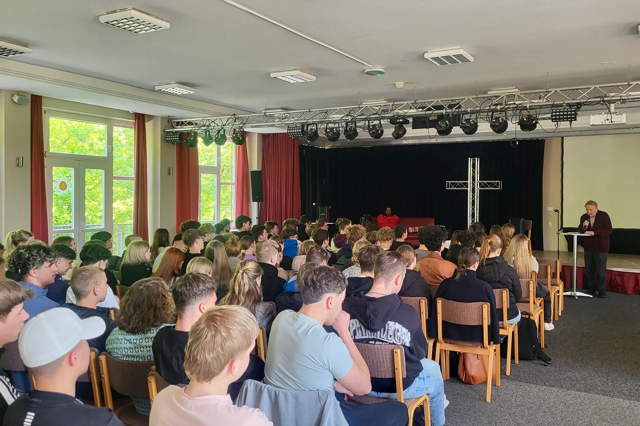 Große Gruppe Schüler sitzt auf Stühlen in einer Aula und lauscht einem Mann in rotem Anzug, der an einem Pult steht. Im Hintergrund ein großes Kreuz und eine dunkle Bühne.