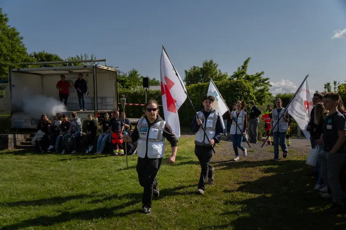 Zwei junge Menschen tragen beim Umzug der 7. Rotkreuzspiele weiße Flaggen und Westen mit dem Logo des Deutschen Roten Kreuzes. Im Hintergrund sind weitere Teilnehmende und ein Fahrzeug zu sehen.