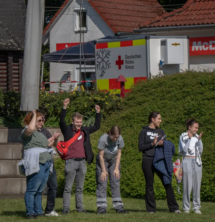 Eine Gruppe junger Menschen steht auf einer Wiese vor einem weißen Zelt des Deutschen Roten Kreuzes und hält die Arme in die Höhe.