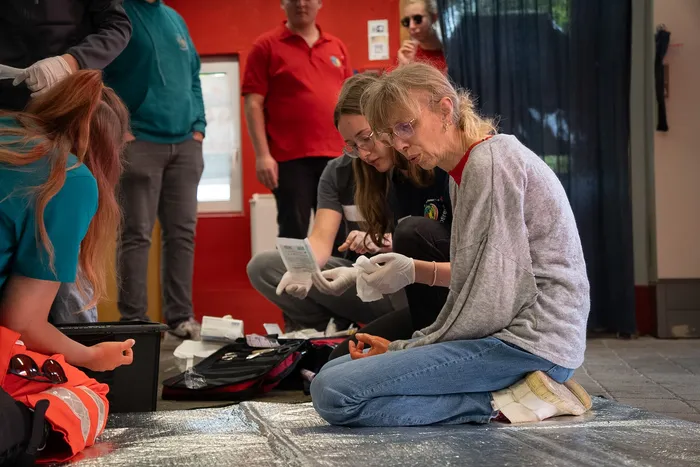 Zwei Sanitäterinnen knien neben einer verletzten Person und versorgen deren Hand. Im Hintergrund weitere Einsatzkräfte und Zuschauer bei den Rotkreuzspielen.