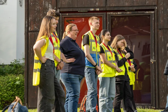 Fünf junge Menschen und eine erwachsene Frau in Warnwesten stehen vor einem hölzernen Gebäude. Die Gruppe scheint auf etwas zu warten oder eine Aufgabe zu besprechen.