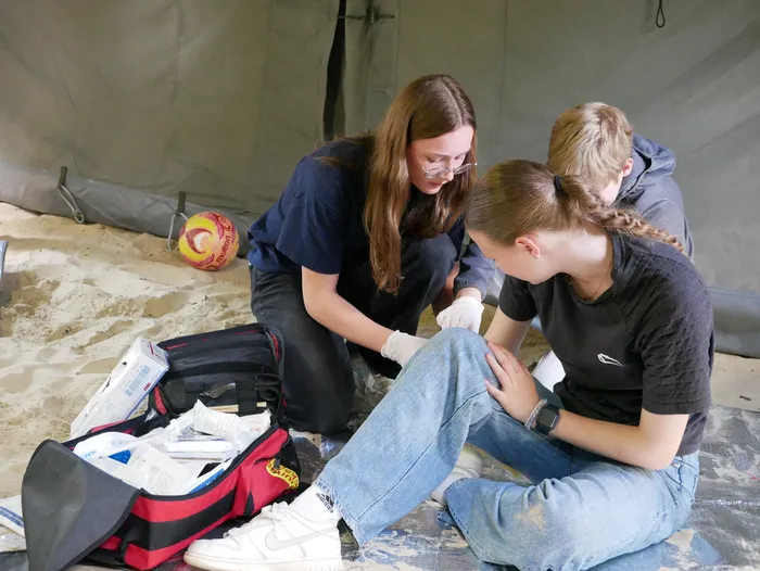 Drei junge Frauen betreuen eine Verletzte am Fuß in einem Zelt. Eine der Helferinnen trägt Handschuhe und untersucht die Wunde, während die anderen beiden aufmerksam zusehen. Ein Erste-Hilfe-Koffer steht neben der Verletzten.
