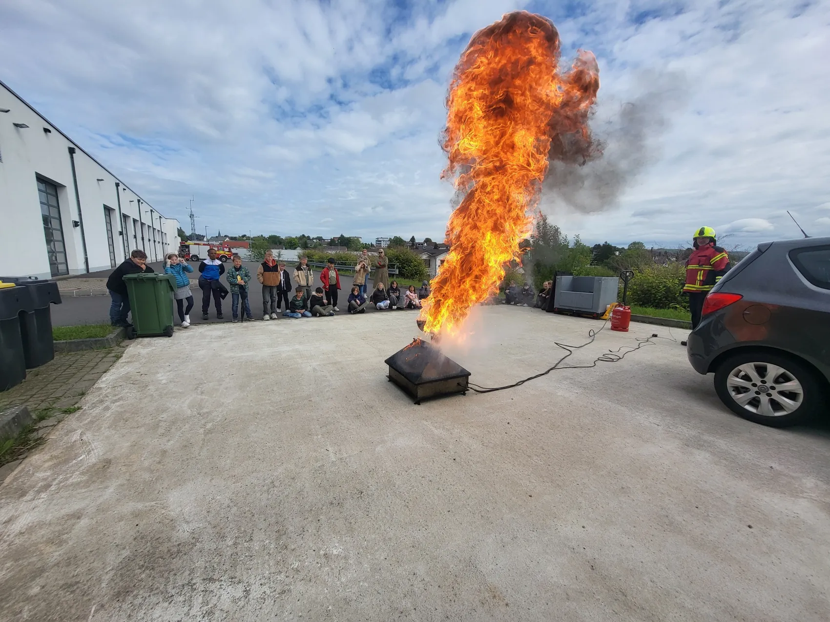 Ein großer Feuerball lodert aus einem Metallbehälter auf einem Pflasterplatz, während eine Gruppe von Kindern und Betreuern zusieht. Ein Feuerwehrmann in voller Montur steht rechts im Bild neben einem Auto und Feuerlöschern.