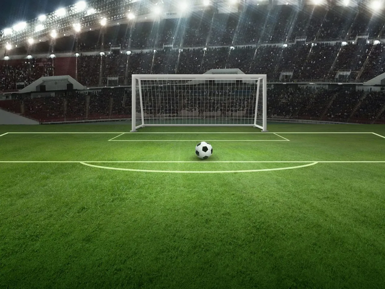 Fußballfeld mit Tor und Ball im Fokus, im Hintergrund ein voll besetztes Stadion bei Nacht.