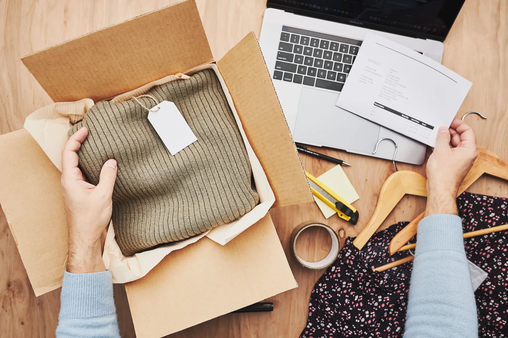 Ausgepackter Pullover mit Etikett neben geöffnetem Karton, Laptop und Zubehör auf Holztisch.