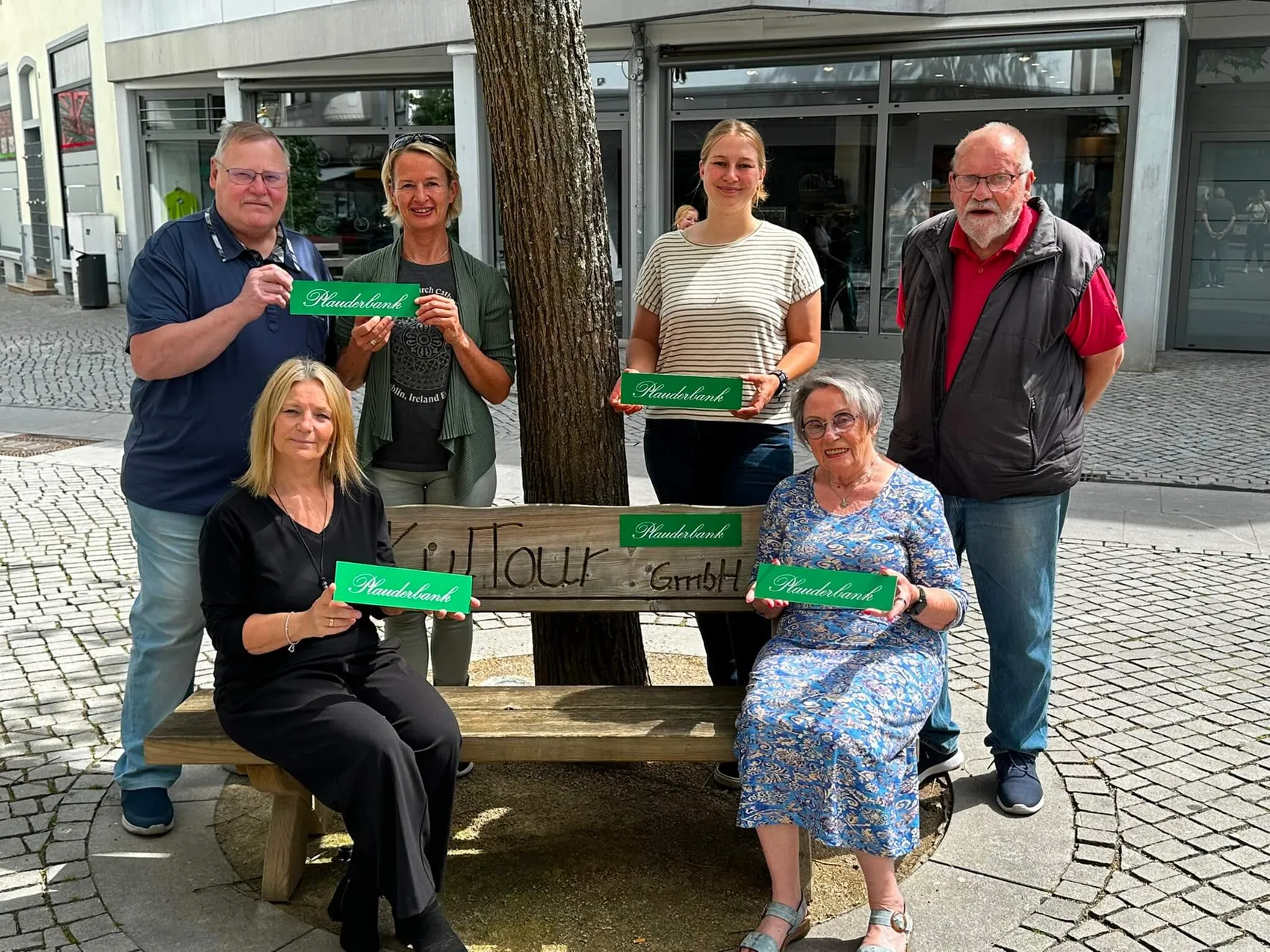 Mehrere Personen halten grüne Schilder mit der Aufschrift „Plauderbank“ vor einer Glasfassade. Sie stehen vor einer neuen Sitzgelegenheit im öffentlichen Raum, die von einem Senioren-Netzwerk aufgestellt wurde.