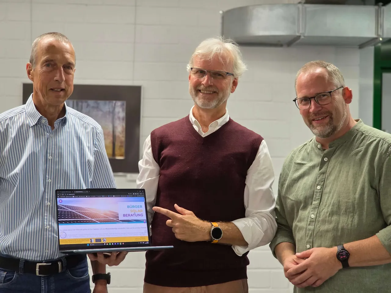 Drei Männer stehen nebeneinander vor einer weißen Wand und zeigen auf einen Laptop. Auf dem Bildschirm ist eine Grafik von Solarmodulen und der Text „Bürger Solar Beratung“ zu sehen.