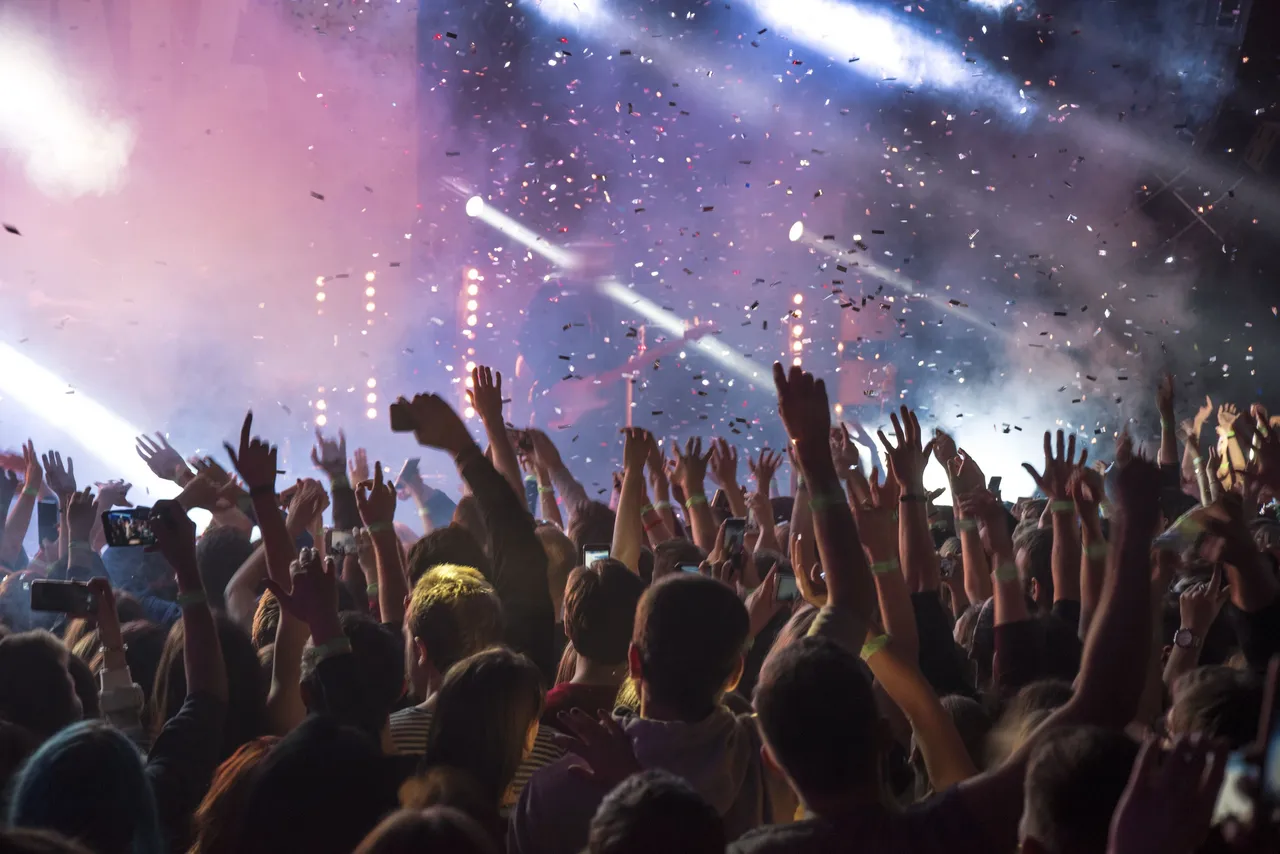 Eine jubelnde Menschenmenge auf einem Konzert oder Festival, viele heben ihre Arme und Smartphones in die Luft. Konfetti fällt von oben.