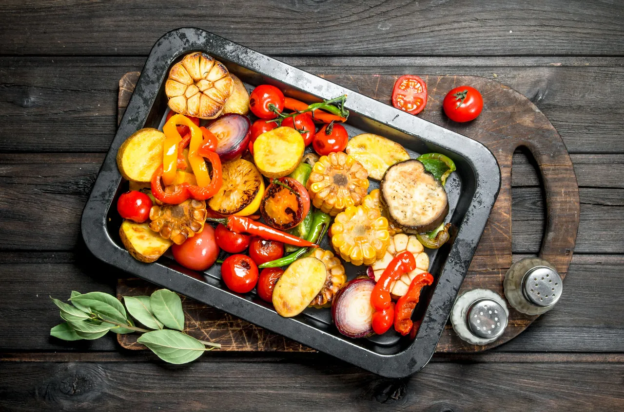 Geröstetes Gemüse in einer dunklen Metallform auf einem Holztisch. Darunter befinden sich frische Tomaten und Kräuter. Salz- und Pfefferstreuer stehen daneben.