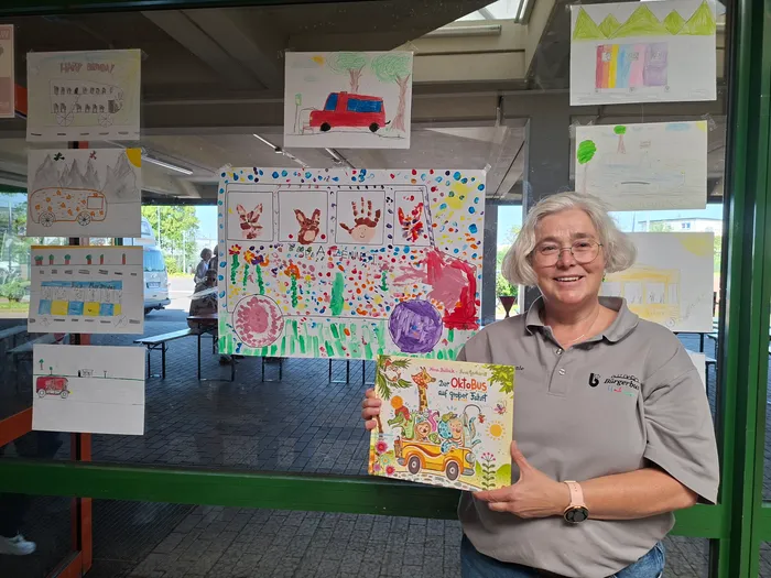 A woman with short gray hair smiles while holding a children's book titled 'Der OktoBus auf großer Fahrt' in front of a display of colorful children's drawings. The drawings are exhibited in the window of a building.