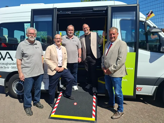 Sechs Männer stehen vor einem barrierefreien Kleinbus mit ausgefahrener Rampe. Sie lächeln in die Kamera und tragen formelle oder legere Kleidung. Im Hintergrund ist eine deutsche Flagge zu sehen.