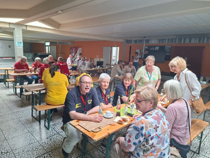 Eine Gruppe von Menschen sitzt an Biertischgarnituren in einer hellen Halle und unterhält sich. Einige tragen Namensschilder und rote Poloshirts mit Aufschrift, andere haben Lanyards um den Hals. Im Vordergrund steht ein Mann in gelbem T-Shirt, der lacht und mit einer Frau spricht.
