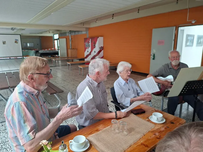 Eine Gruppe älterer Menschen sitzt an einem Holztisch und hält Blätter mit Text in den Händen, während eine Frau singt und ein Mann eine Gitarre spielt. Im Hintergrund sind weitere Personen und ein orangefarbener Raum zu sehen.