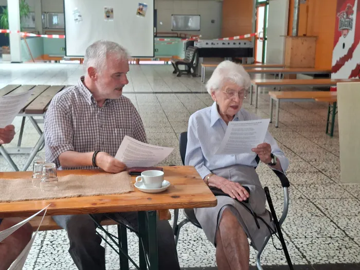 Zwei ältere Menschen, ein Mann und eine Frau, sitzen an einem Holztisch und lesen Dokumente. Sie scheinen sich zu unterhalten, während auf dem Tisch eine Tasse und ein Glas Wasser stehen.
