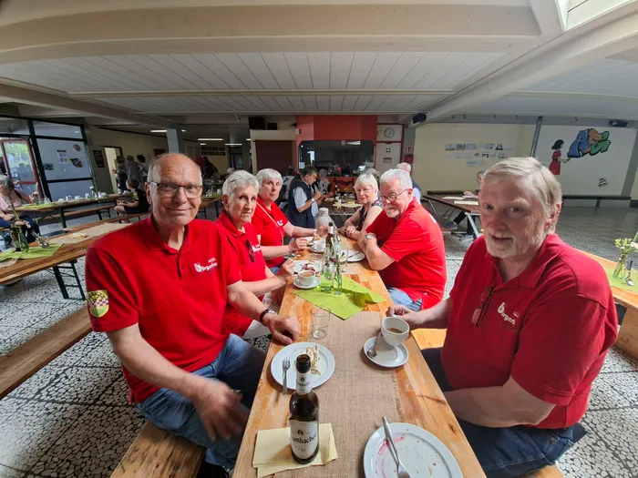 Eine Gruppe von Menschen sitzt an einem Holztisch in einem hellen Raum und isst. Die meisten tragen rote Poloshirts mit dem Aufdruck 