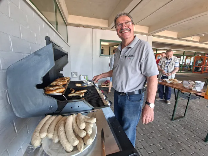Ein Mann mit Brille und Polohemd steht lächelnd vor einem Grill mit Bratwürsten. Im Hintergrund weitere Personen und ein Tisch mit Essen.