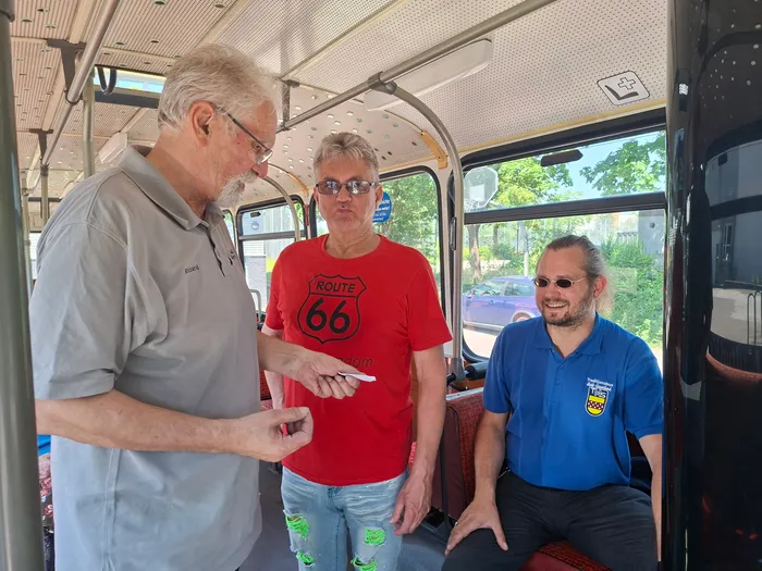 Drei Männer stehen in einem Oldtimerbus und unterhalten sich. Der Mann rechts trägt ein blaues Poloshirt mit einem Logo, der Mann in der Mitte ein rotes T-Shirt mit der Aufschrift 