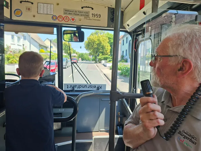 Ein Mann mit grauem Haar und Bart hält ein Funkgerät in einem Bus, während ein jüngerer Mann mit dem Rücken zum Betrachter am Lenkrad sitzt. Durch die Windschutzscheibe ist eine Straße mit Häusern zu sehen.