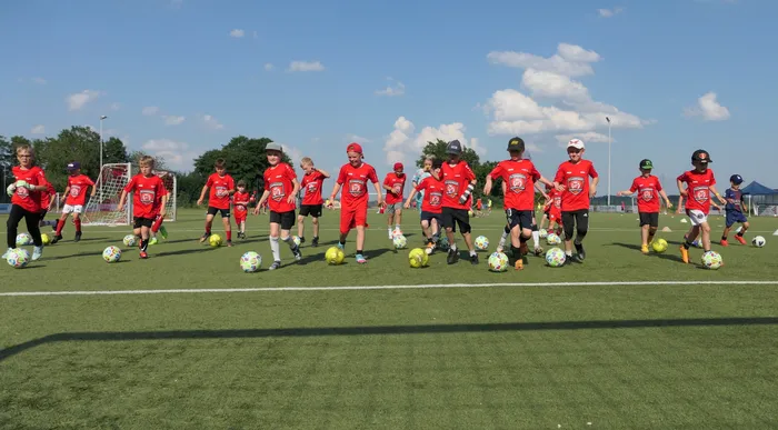 Eine Gruppe Jugendlicher in roten Trikots trainiert auf einem grünen Fußballfeld. Jeder Spieler hat einen Ball vor sich und bereitet sich auf eine Übung vor.