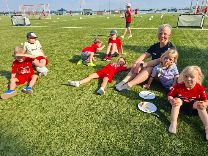 Eine Gruppe von Kindern in roten Trikots und eine erwachsene Person sitzen auf dem grünen Rasen eines Fußballfeldes und essen Kuchen. Im Hintergrund sind weitere Kinder beim Fußballtraining zu sehen.