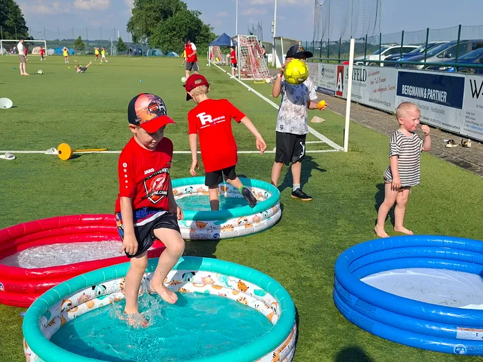 Kinder planschen und spielen an einem sonnigen Tag in kleinen Pools auf einem Rasenplatz, während im Hintergrund ein Fußballspiel stattfindet.