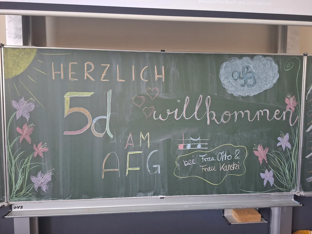 Herzlich willkommen zur 5d am AFG – so steht es in bunter Kreide auf der grünen Tafel, umrahmt von Schmetterlingen und Blumen. Darunter eine Zeichnung eines Musikinstruments und die Namen der Lehrerinnen Frau Otto und Frau Karstel.