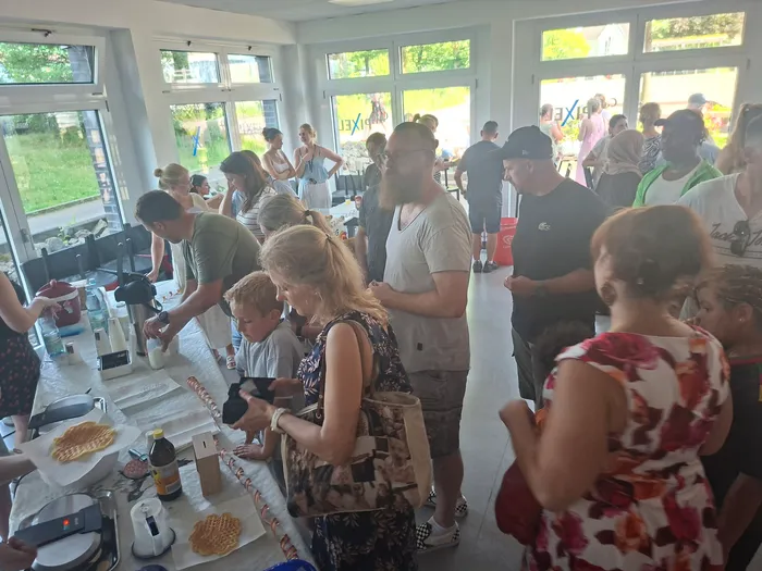 Menschen stehen und arbeiten an Tischen mit Lebensmitteln, vermutlich bei der Zubereitung von Speisen. Im Hintergrund sind große Fenster zu einem grünen Außenbereich zu sehen.