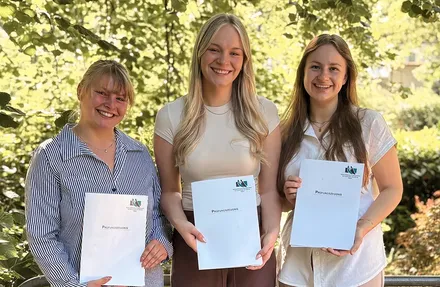 Drei junge Frauen halten jeweils ein Prüfungszeugnis in der Hand und lächeln in die Kamera. Im Hintergrund ist ein verschwommener Park zu sehen.
