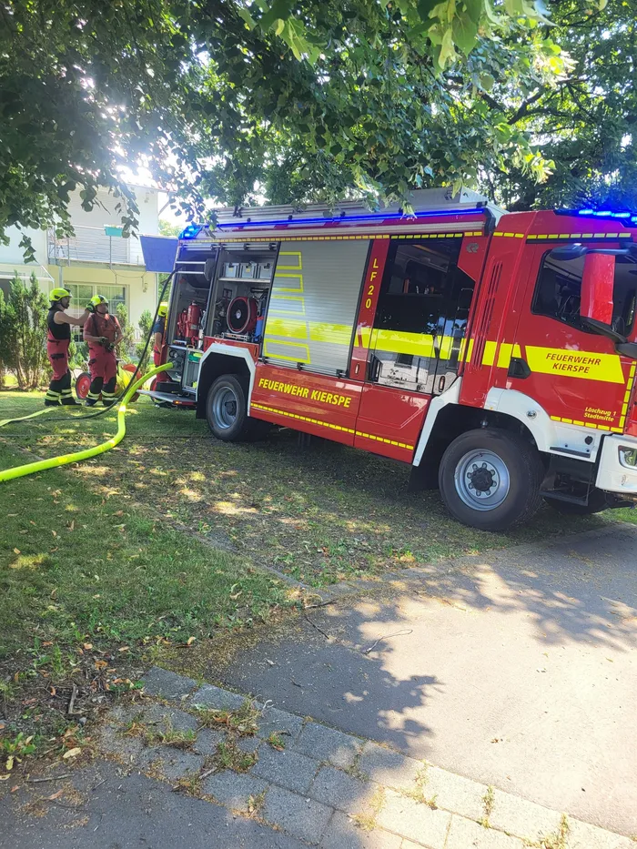 Roter Feuerwehrauto mit der Aufschrift „Feuerwehr Kierspe“ steht auf einer Straße, während ein Feuerwehrmann einen Schlauch ausrollt. Im Hintergrund sind weitere Einsatzkräfte und ein Gebäude zu sehen.