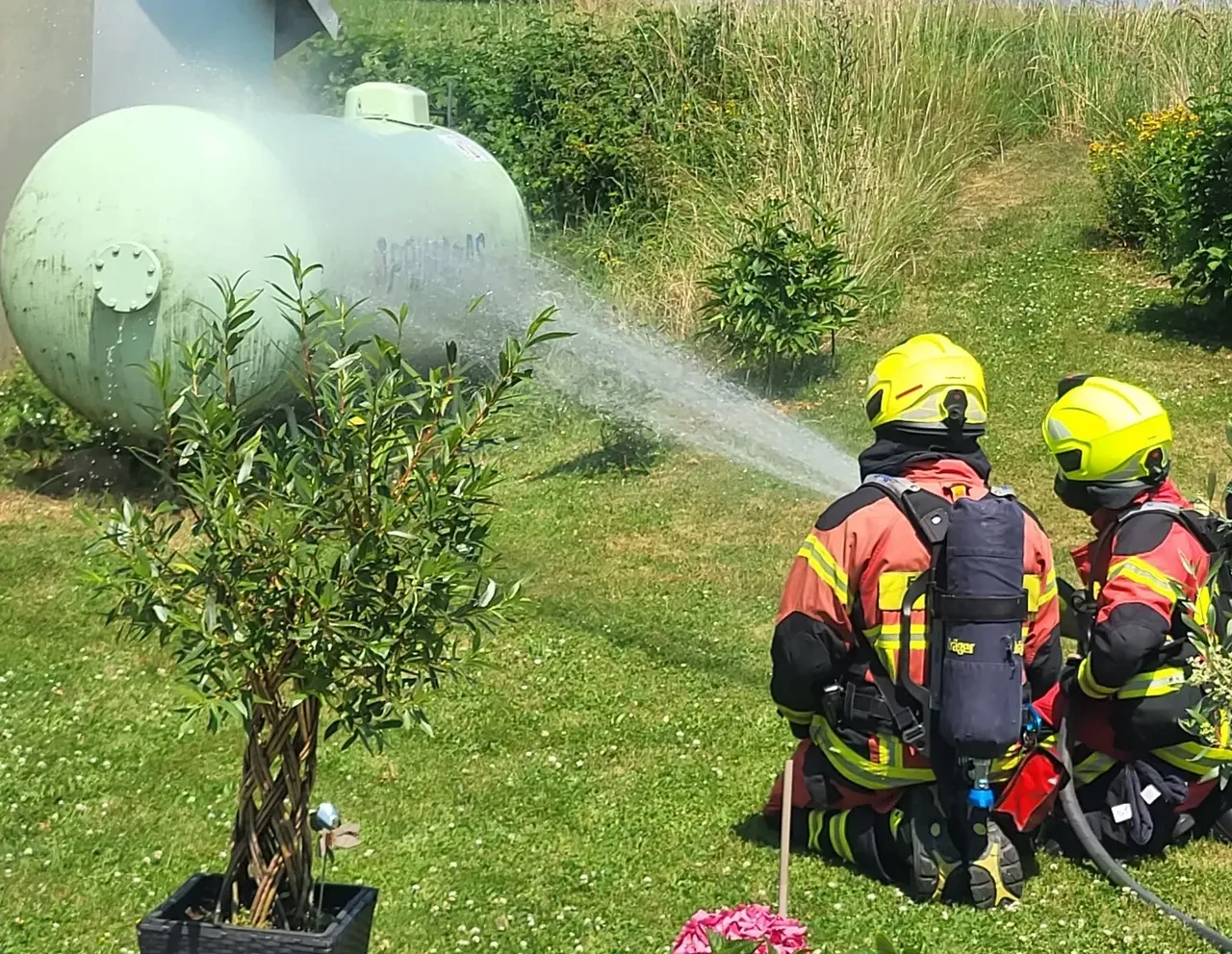 Zwei Feuerwehrleute kühlen einen großen, zylindrischen Gastank mit Wasser ab. Im Vordergrund stehen Kübelpflanzen auf einer Terrasse.