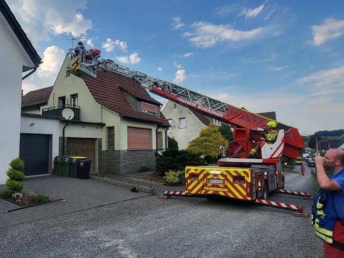 Feuerwehrleute bedienen eine ausgefahrene Drehleiter an einem Wohnhaus. Im Hintergrund sind weitere Einsatzfahrzeuge und ein bewölkter Himmel zu sehen.