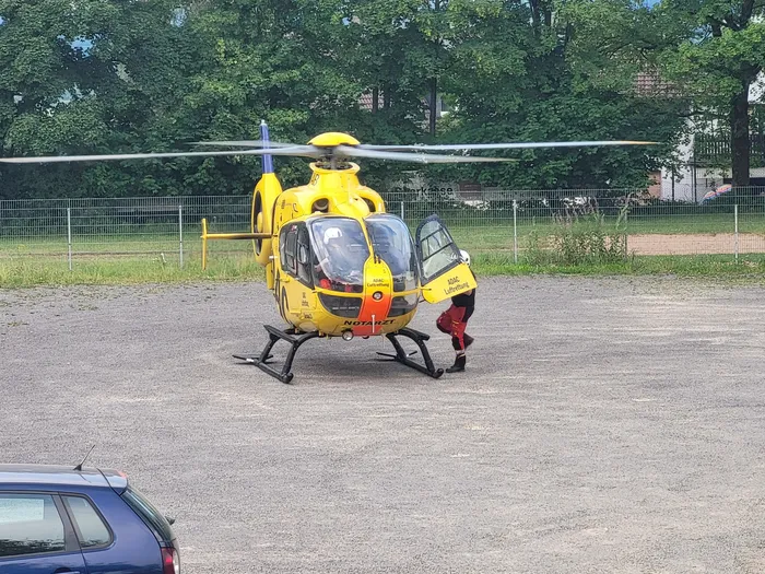 Gelber Rettungshubschrauber steht auf einer Wiese, während eine Person in roter Einsatzkleidung in seine Nähe geht. Im Hintergrund sind Bäume und ein Zaun zu sehen.