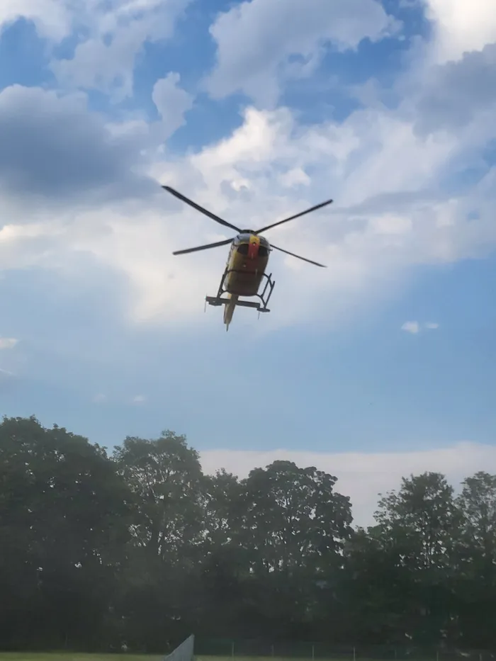 Ein gelb-roter Rettungshubschrauber fliegt vor einem bewölkten Himmel über Baumwipfeln. Im Hintergrund ist eine Grünfläche zu erkennen.