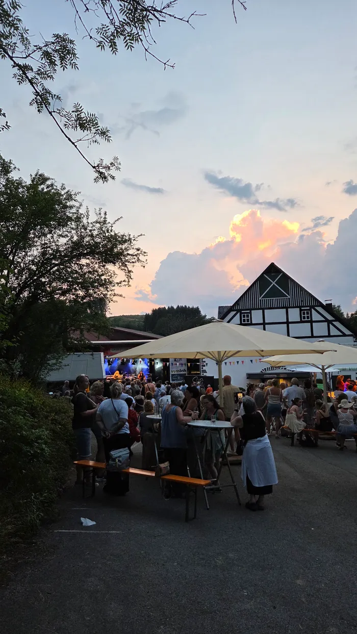 Das Bild zeigt eine Open-Air-Veranstaltung bei Sonnenuntergang. Menschen sitzen an Biertischbänken vor einer Bühne, auf der eine Band spielt, vor dem Hintergrund eines traditionellen Fachwerkhouses.